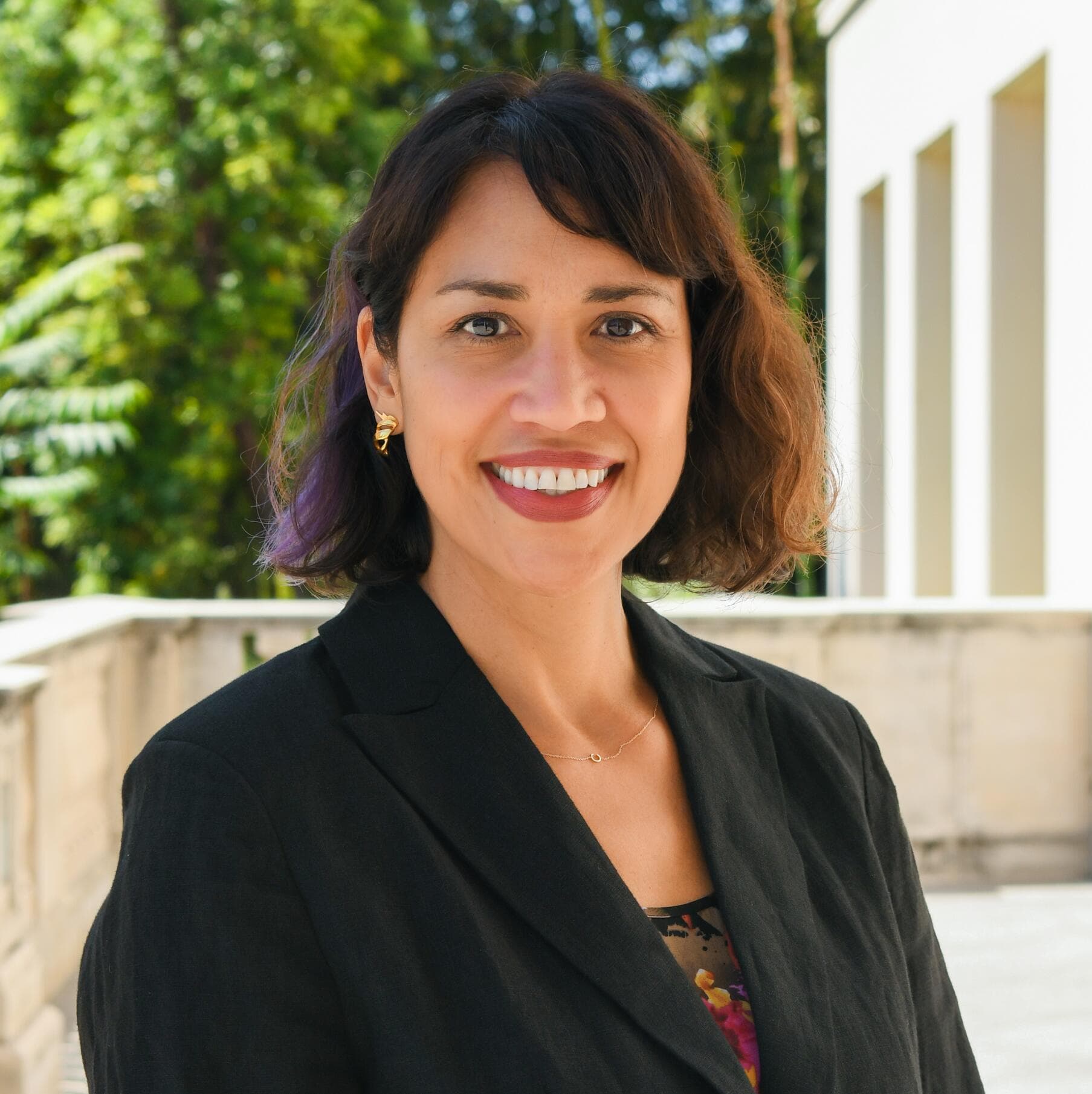 a person with bobbed hair wears a black blazer and smiles at the camera while standing outdoors.