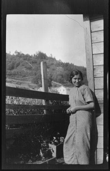 An unidentified Native American young woman standing next to a wooden building and fence