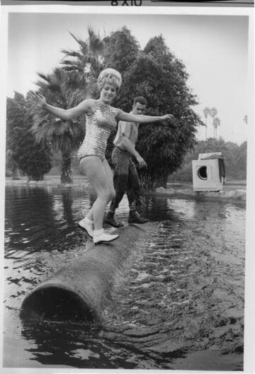 Photo of a woman in a bathing suit, and a lumberjack rolling a log in a pond with an electric dryer on the bank in the background.                   '