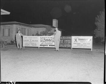 Two men standing on front lawn of new home next to signs for Adequate Wiring, Lighting, Remote systems, and electric appliances